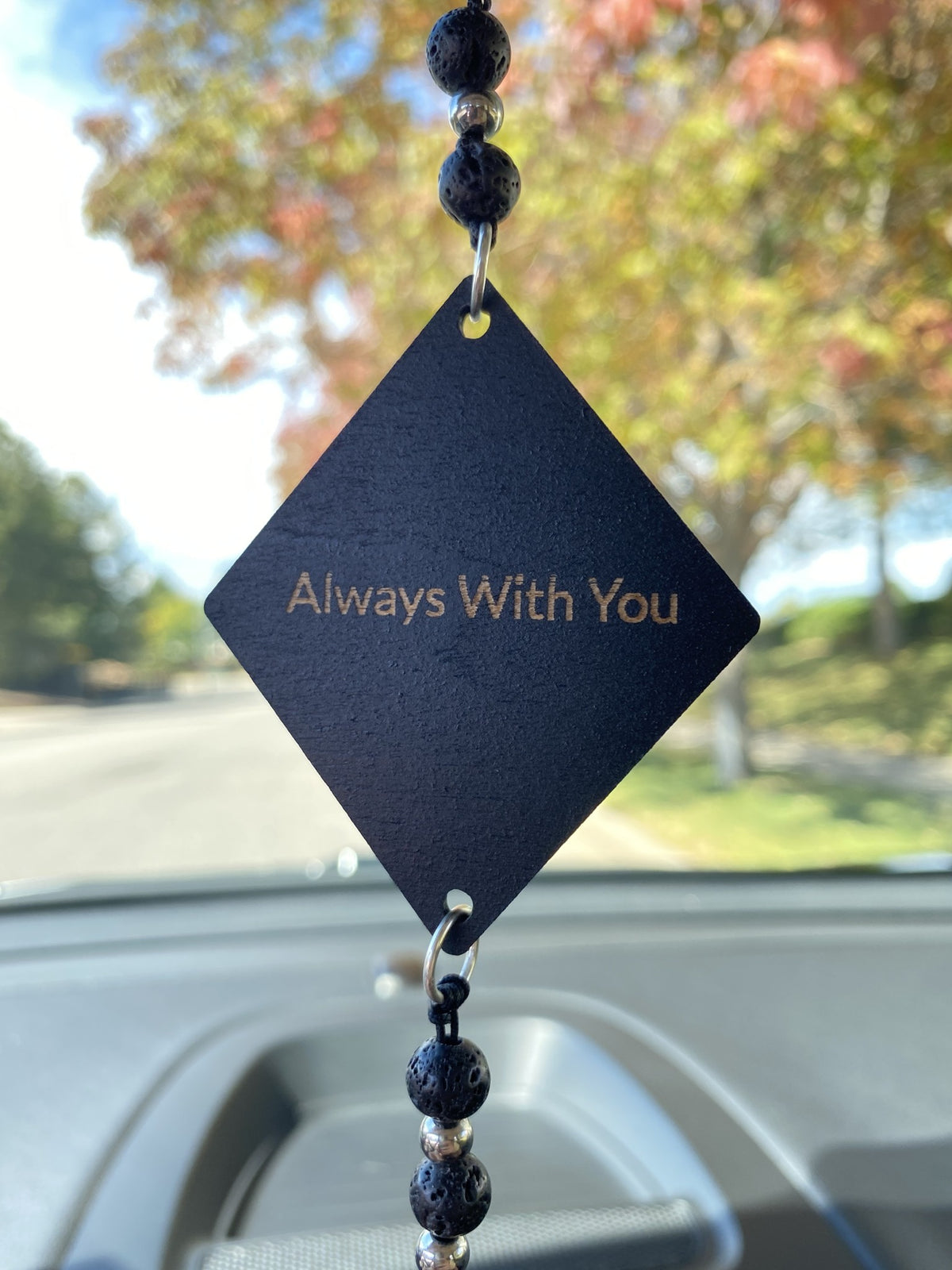 A black diamond-shaped car air freshener with 'Always With You' printed in gold, hanging in a car window with blurred trees outside.