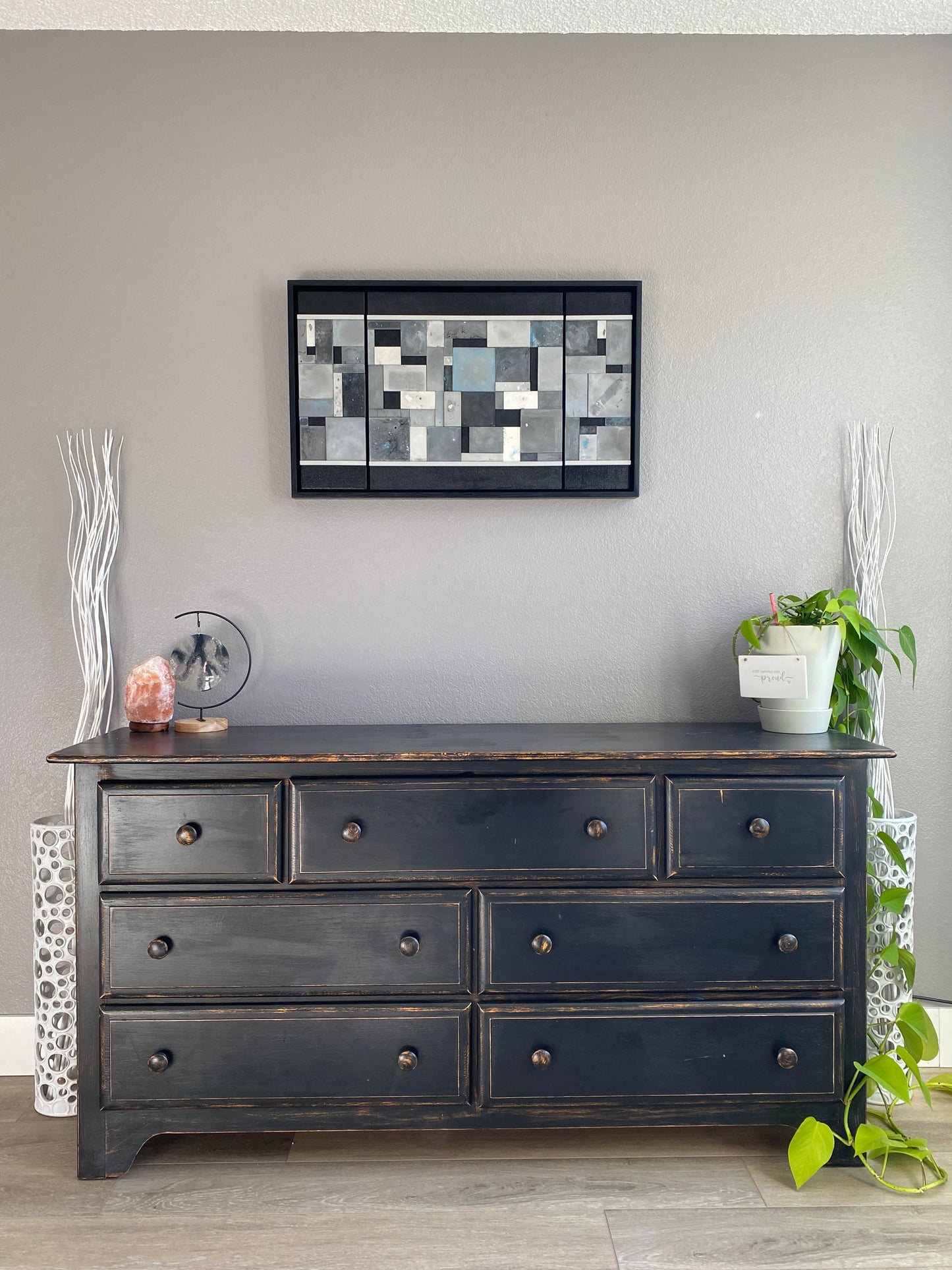 Dark wooden cabinet with multiple drawers against a gray wall, featuring a decorative art piece and plant.