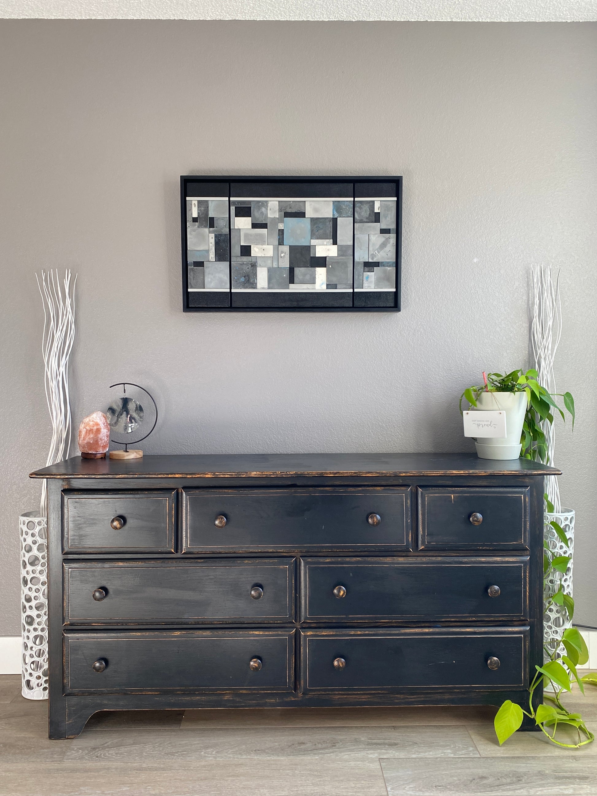 Dark wooden cabinet with multiple drawers against a gray wall, featuring a decorative art piece and plant.