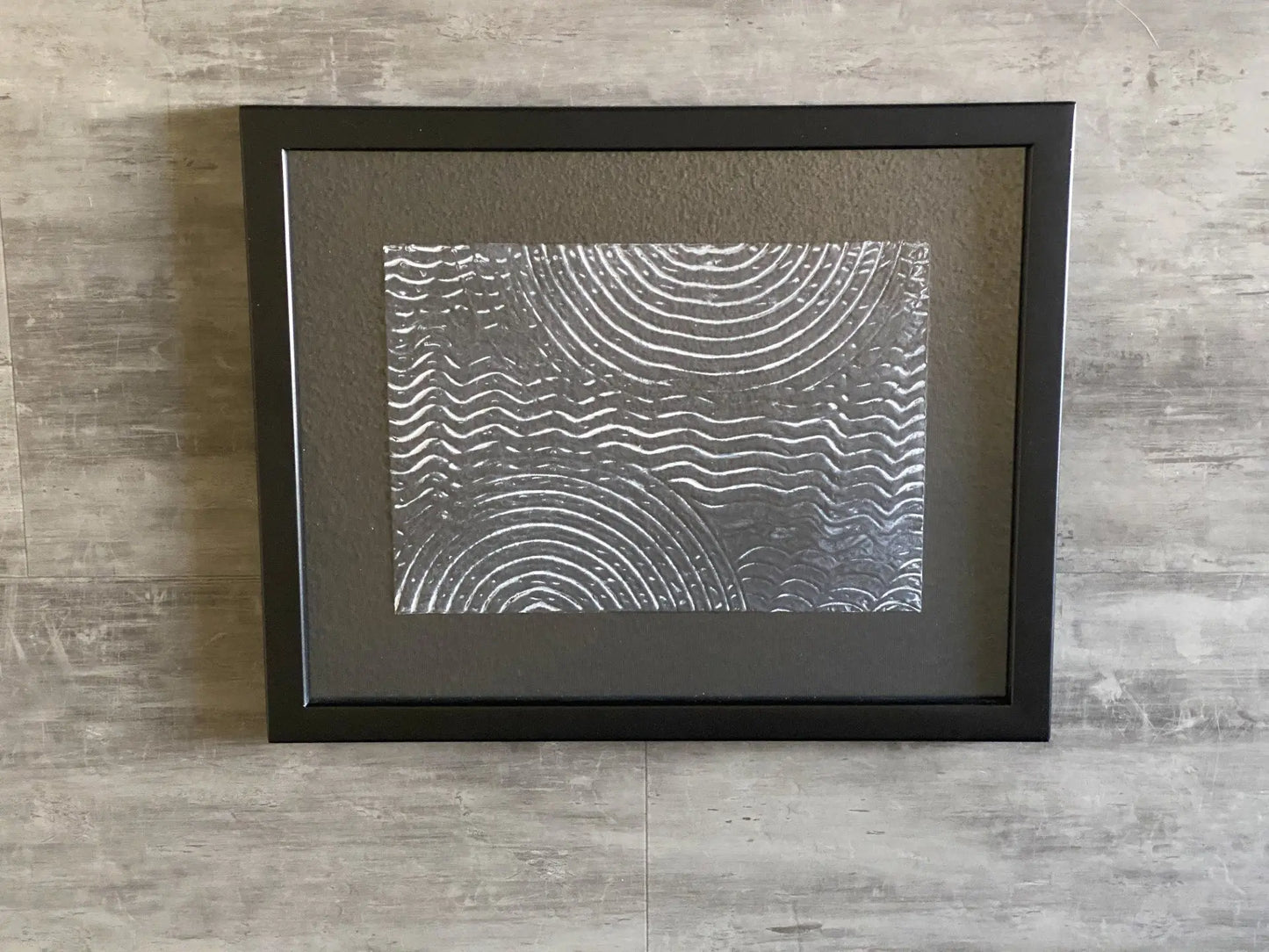 Framed artwork featuring textured acrylic resin with concentric and wavelike patterns on a reflective surface.