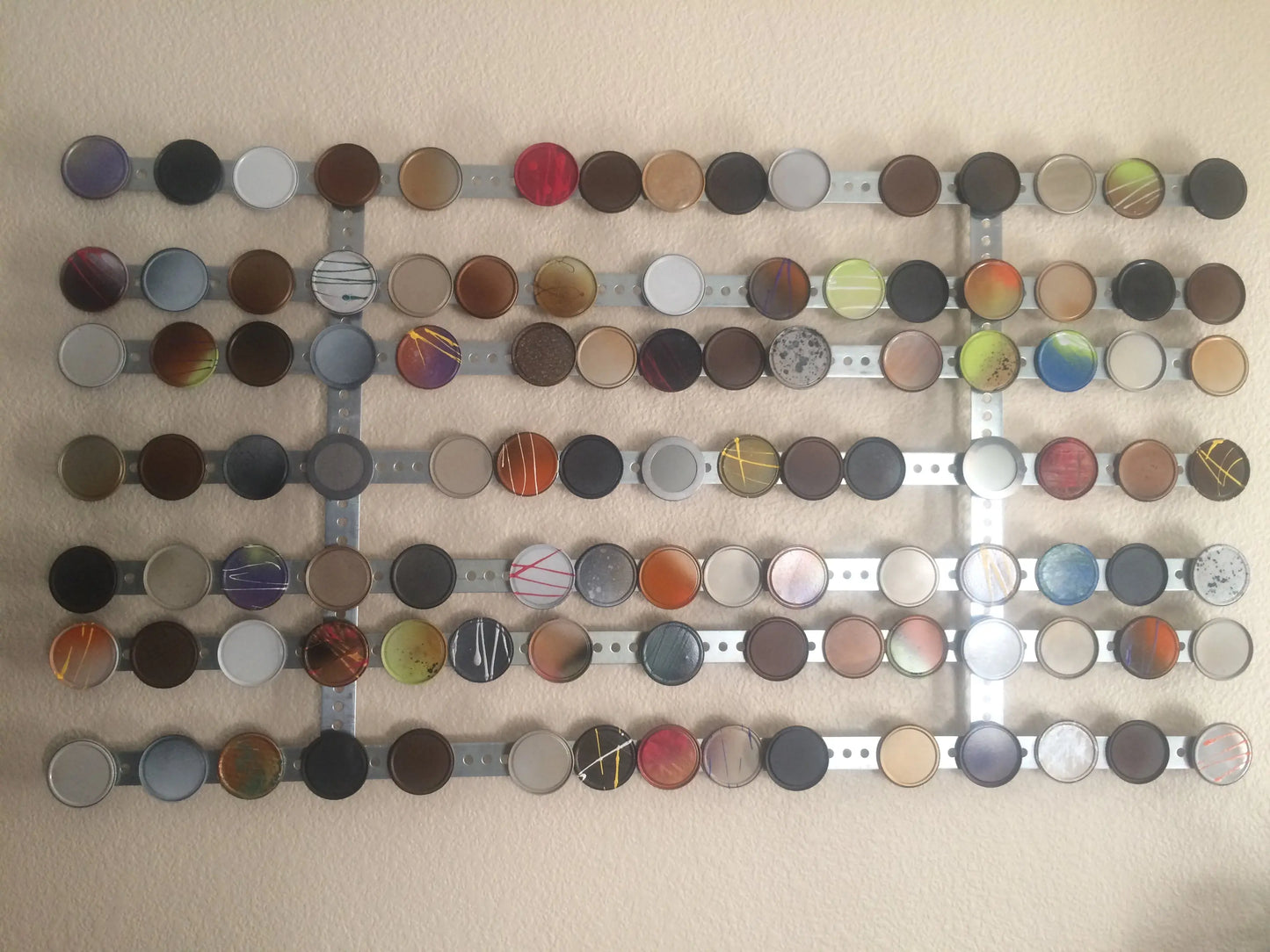 Wall‑mounted mixed‑media artwork featuring painted circular forms made from metal and repurposed juice lids arranged on horizontal metal bars.