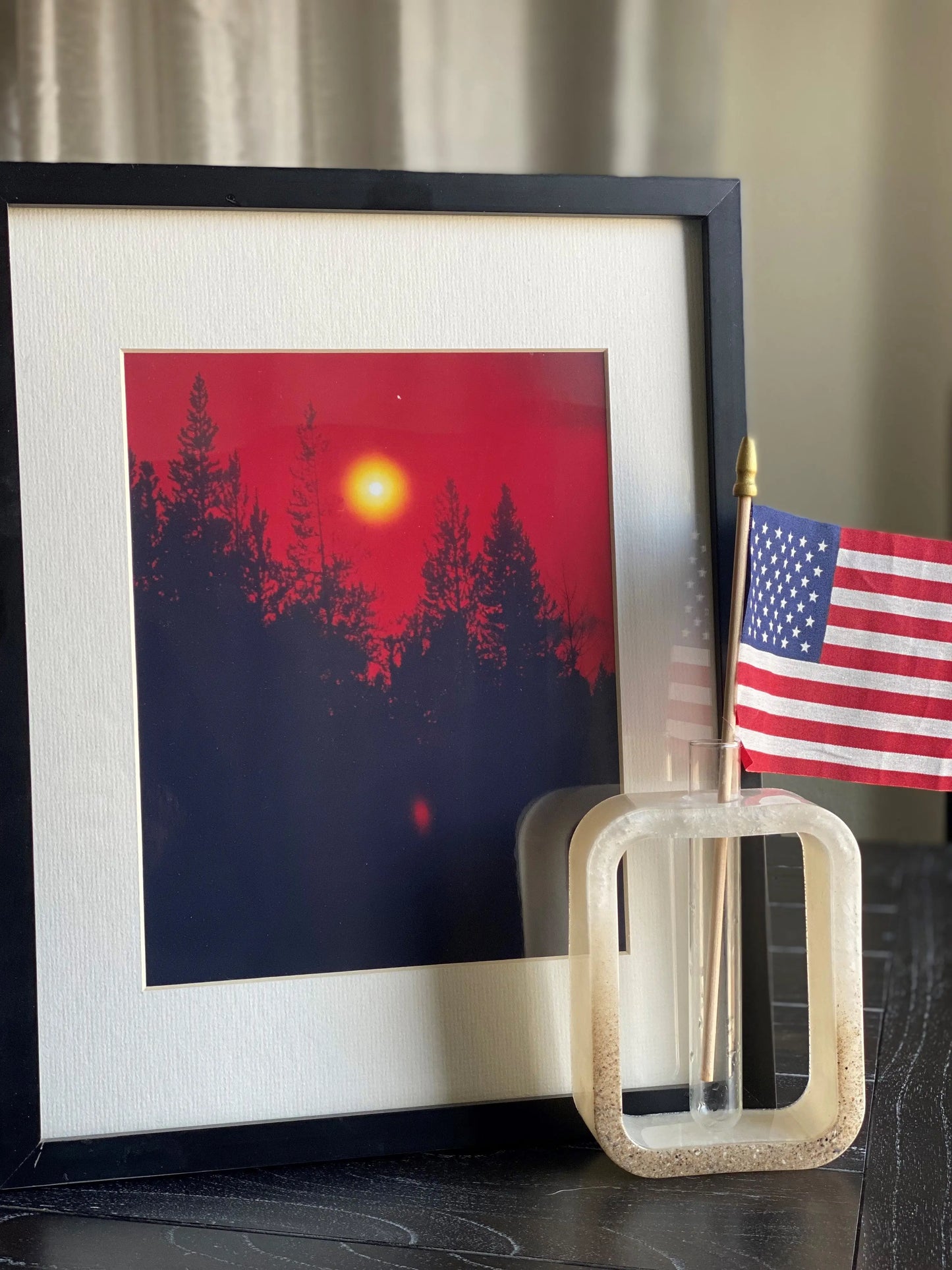 Framed artwork with a red sunset over trees and an American flag on a stand.