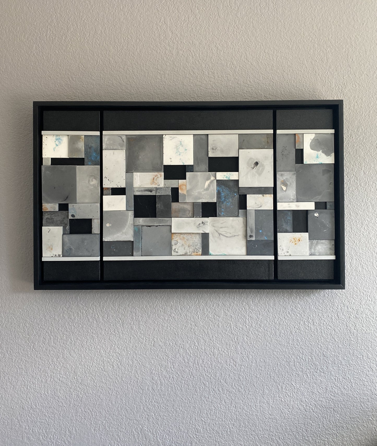 Abstract wall art with geometric shapes in a black frame on a plain wall.