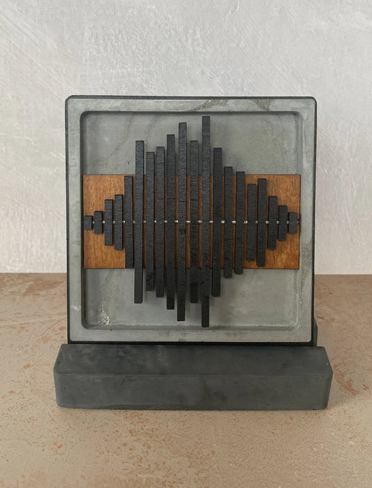 A square abstract wall sculpture with a gray stone or concrete frame and base, featuring a central arrangement of black rectangular bars over warm brown wood panels, set against a textured off-white wall and beige floor.