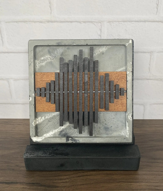 A square abstract wall art sculpture on a dark rectangular base, featuring layered metallic gray bars over a warm brown wood panel, set against a white brick wall and wooden tabletop.