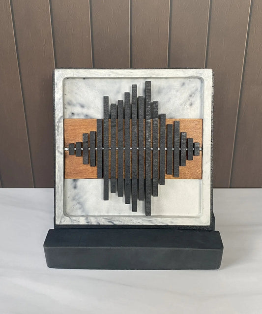 A square abstract wall art sculpture featuring a white and gray marble frame, a central black metallic soundwave-like structure, two horizontal brown wood panels, and a black rectangular base, displayed against a brown paneled wall on a white surface.