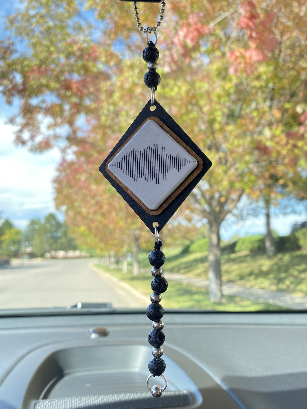 A diamond-shaped car windshield ornament hanging in a car, featuring a black wood frame, a white center with a black soundwave graphic, and a beaded black and silver tassel.