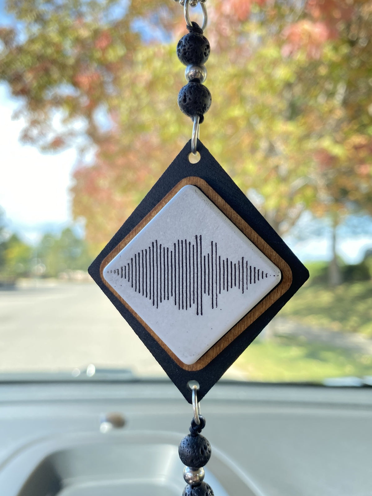 A diamond-shaped car ornament hanging in a car window. It features a black outer frame, a light wood inner border, a white center with a black soundwave graphic, and dark beaded tassels.