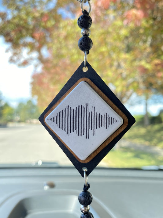 A diamond-shaped car ornament hanging in a car window. It features a black outer frame, a light wood inner border, a white center with a black soundwave graphic, and dark beaded tassels.