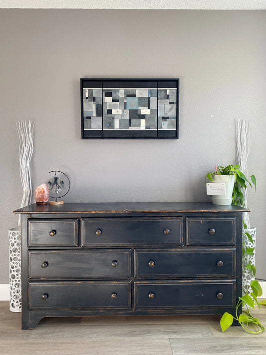 Dark wooden cabinet with multiple drawers against a gray wall, featuring a decorative art piece and plant.