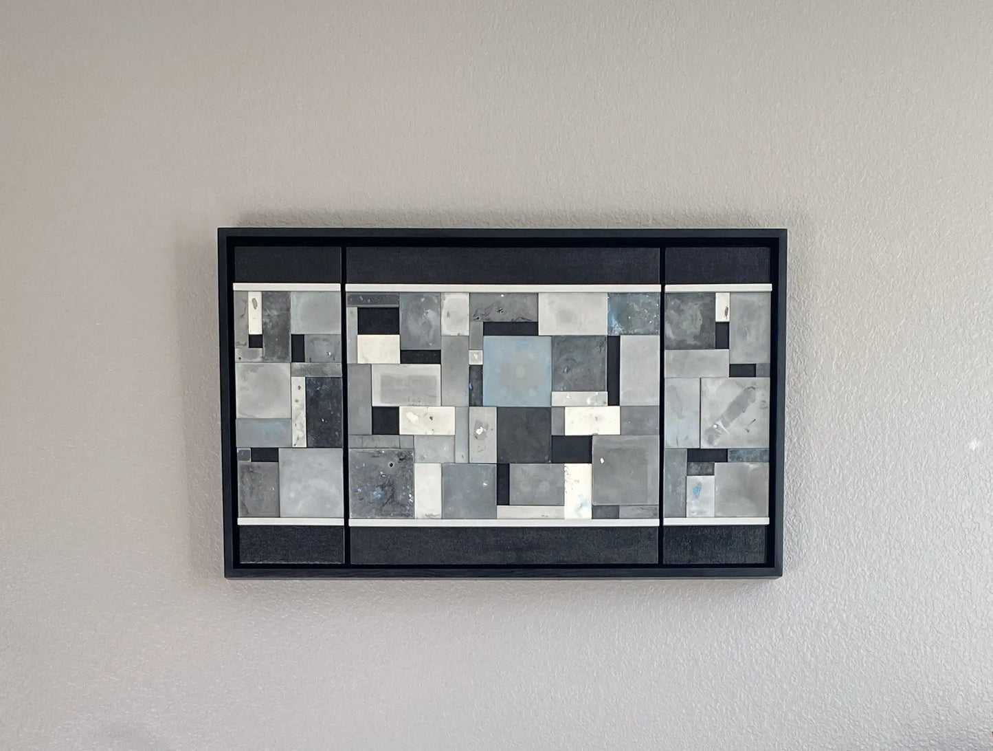 Abstract art piece with geometric shapes in black, white, blue, and gray on a gray wall.