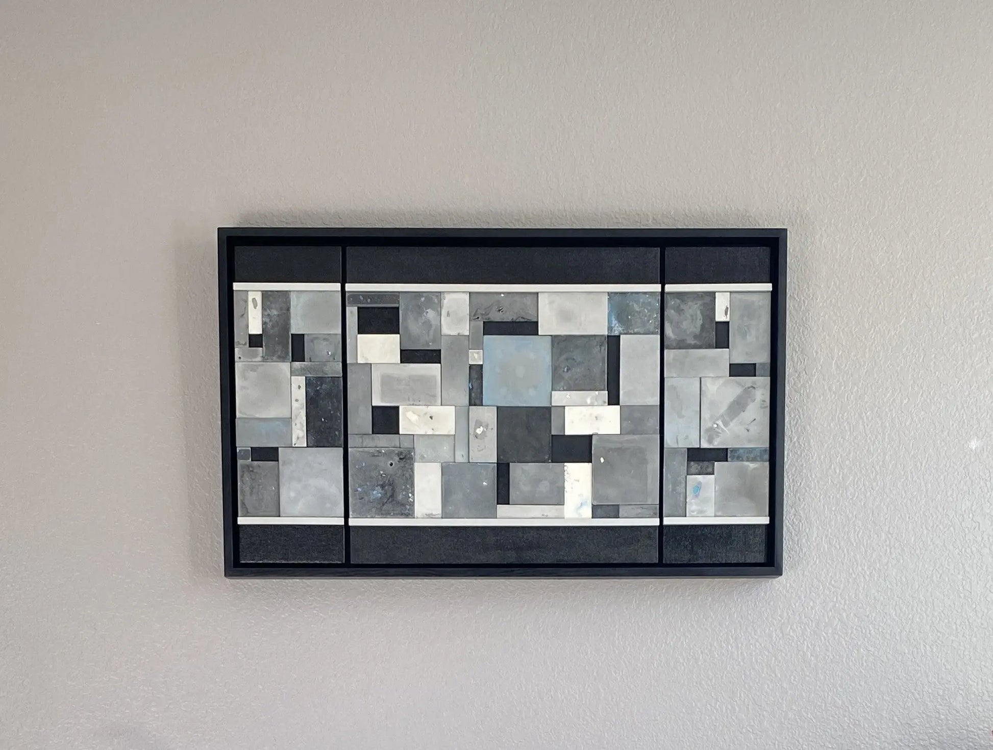 Abstract art piece with geometric shapes in black, white, blue, and gray on a gray wall.