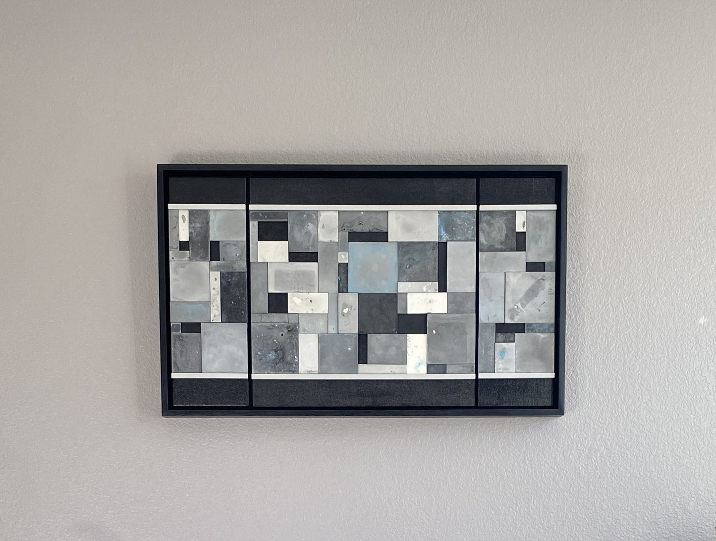 Abstract art piece with geometric shapes in black, white, blue, and gray on a gray wall.