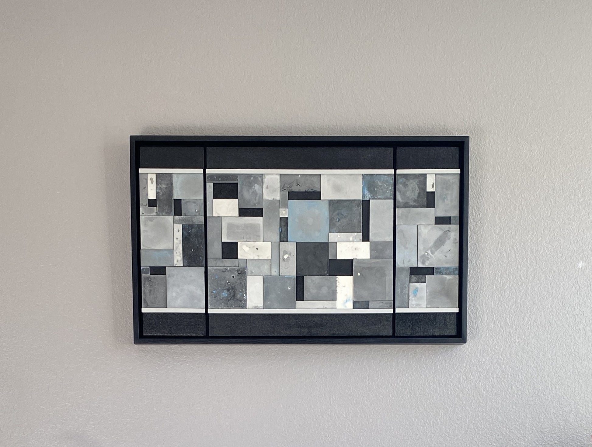 Abstract art piece with geometric shapes in black, white, blue, and gray on a gray wall.