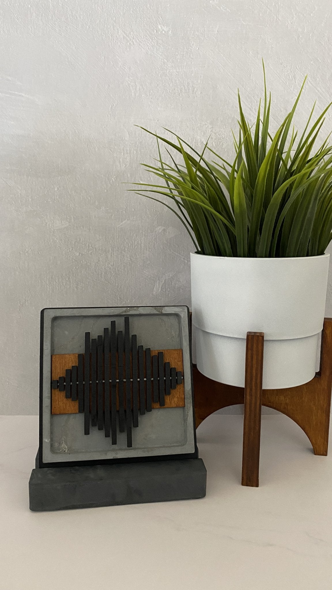 A square decorative black geometric soundwave design on a gray face, displayed next to a potted green plant on a white and wood stand against a textured light gray wall.