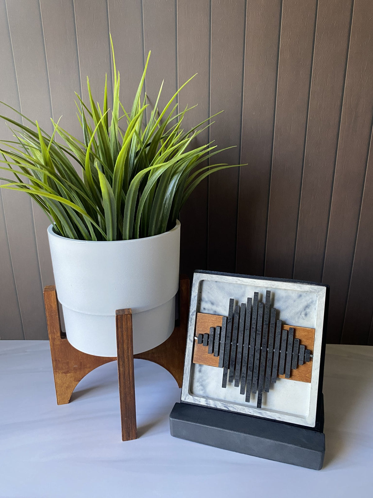 A decorative geometric wall art piece with dark gray slats and wood accents displayed next to a potted green plant on a wooden stand against a brown paneled wall.