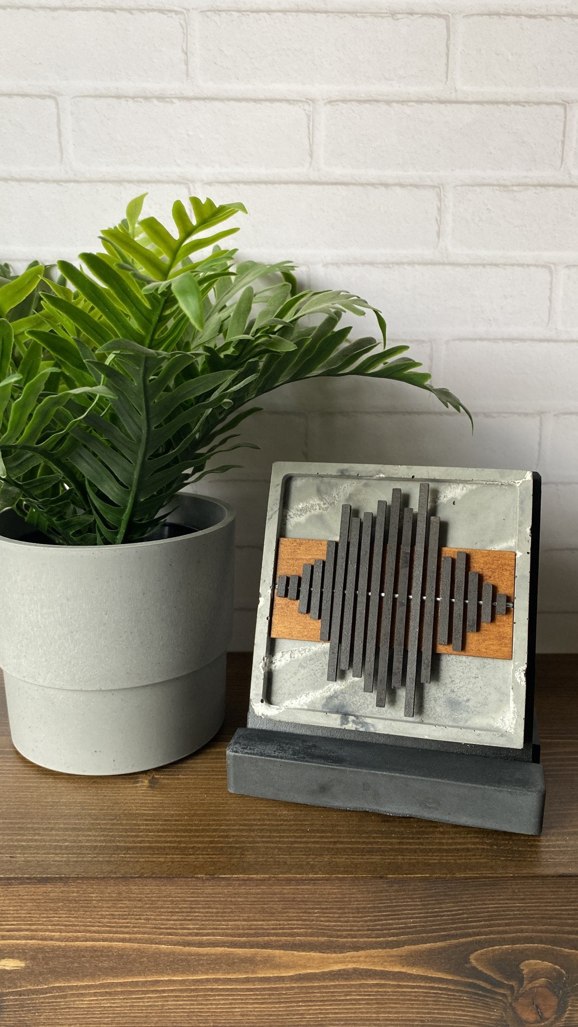 A square decorative sound wave sculpture with a gray marble-look face, dark gray vertical slats, and orange wood accents, displayed on a dark base on a wooden table next to a potted green plant against a white brick wall.