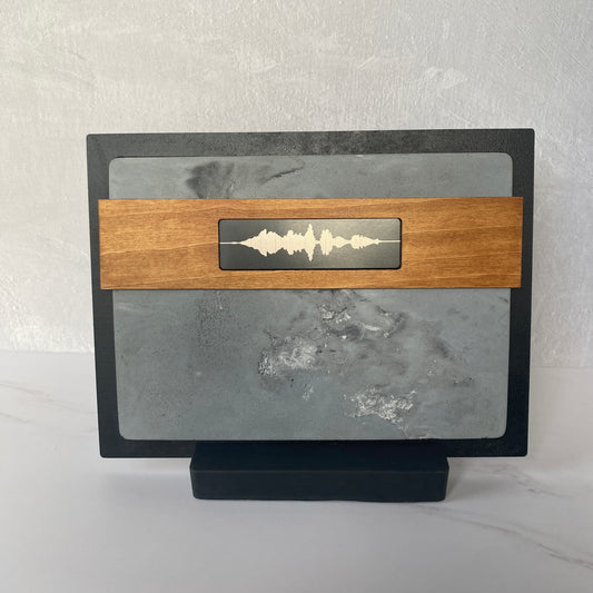 A rectangular soundwave plaque or decorative board on a small black stand, featuring a gray stone-like center with a horizontal wooden band across the middle and a white soundwave motif in the center.
