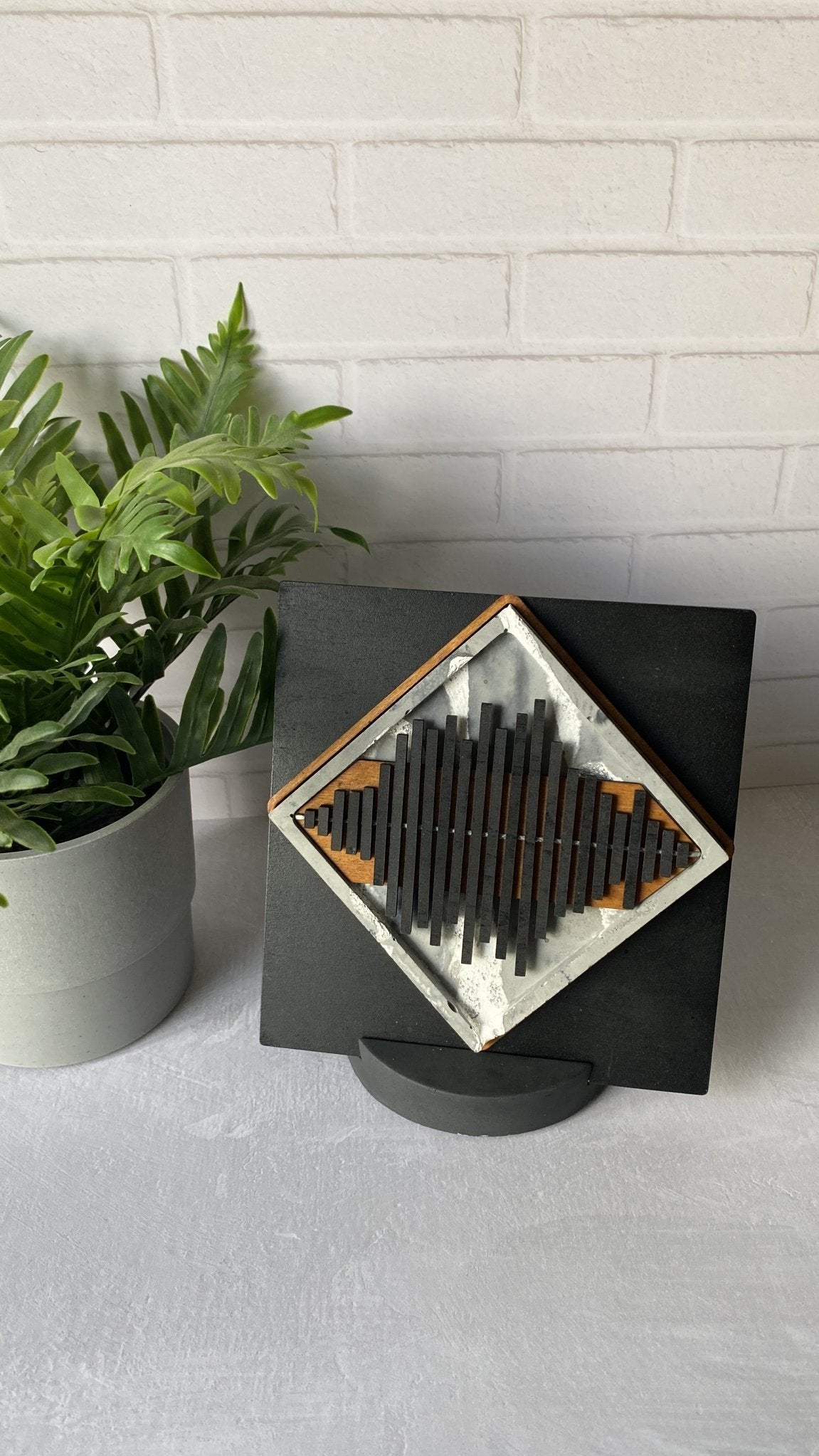 A square black wall art piece with a diamond-shaped central design made of dark vertical bars on a light background, displayed on a stand against a white brick wall next to a potted green plant.