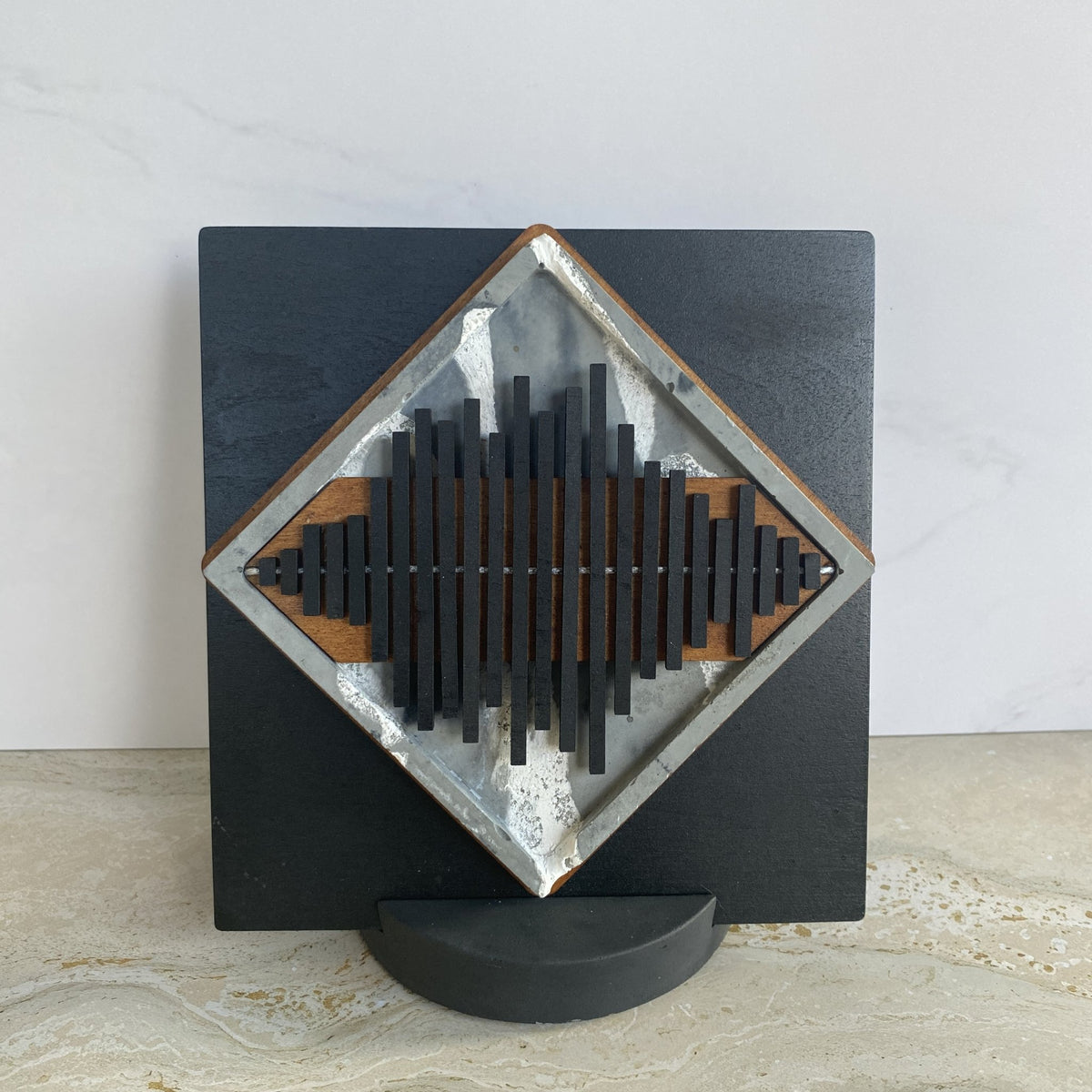 A square black wall-mounted art piece with a diamond-shaped inset featuring a stylized soundwave design in black, silver, and brown wood tones, displayed on a small black stand against a light gray wall and marble surface.