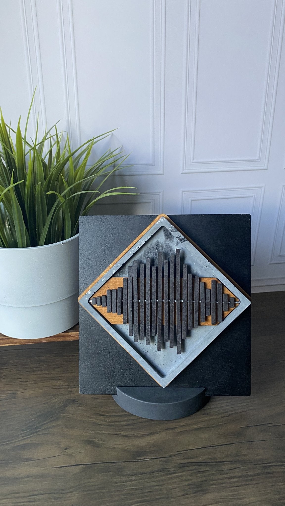 A square black decorative plaque or wall art piece with a diamond-shaped inset featuring vertical dark rods and light wood accents, displayed on a small round stand on a wooden floor next to a potted green plant.