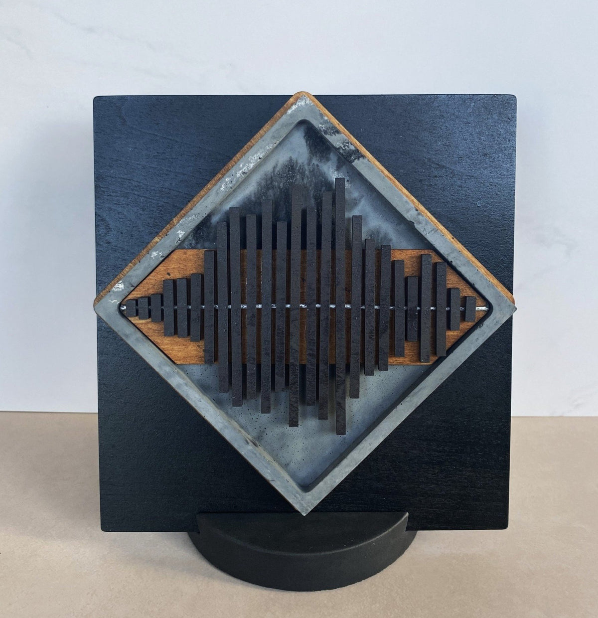 A square black wooden wall art piece with a diamond-shaped inset featuring vertical dark slats over a gray and black textured background, mounted on a black curved base against a light wall and beige floor.
