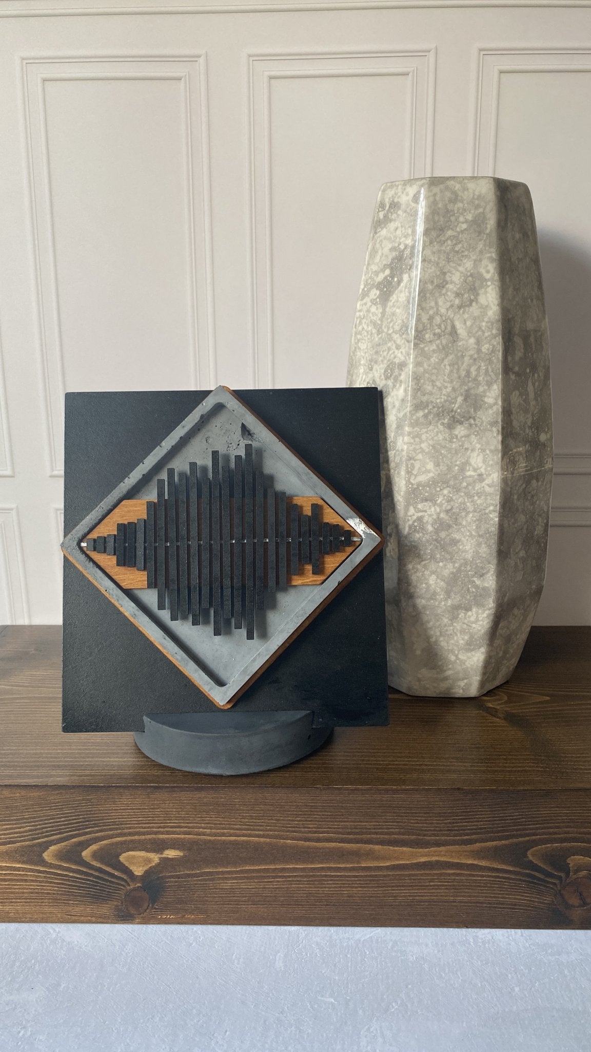 A square black decorative plaque with a diamond-shaped geometric soundwave design in black, gray, and orange, displayed on a round stand next to a tall textured stone vase on a wooden table.