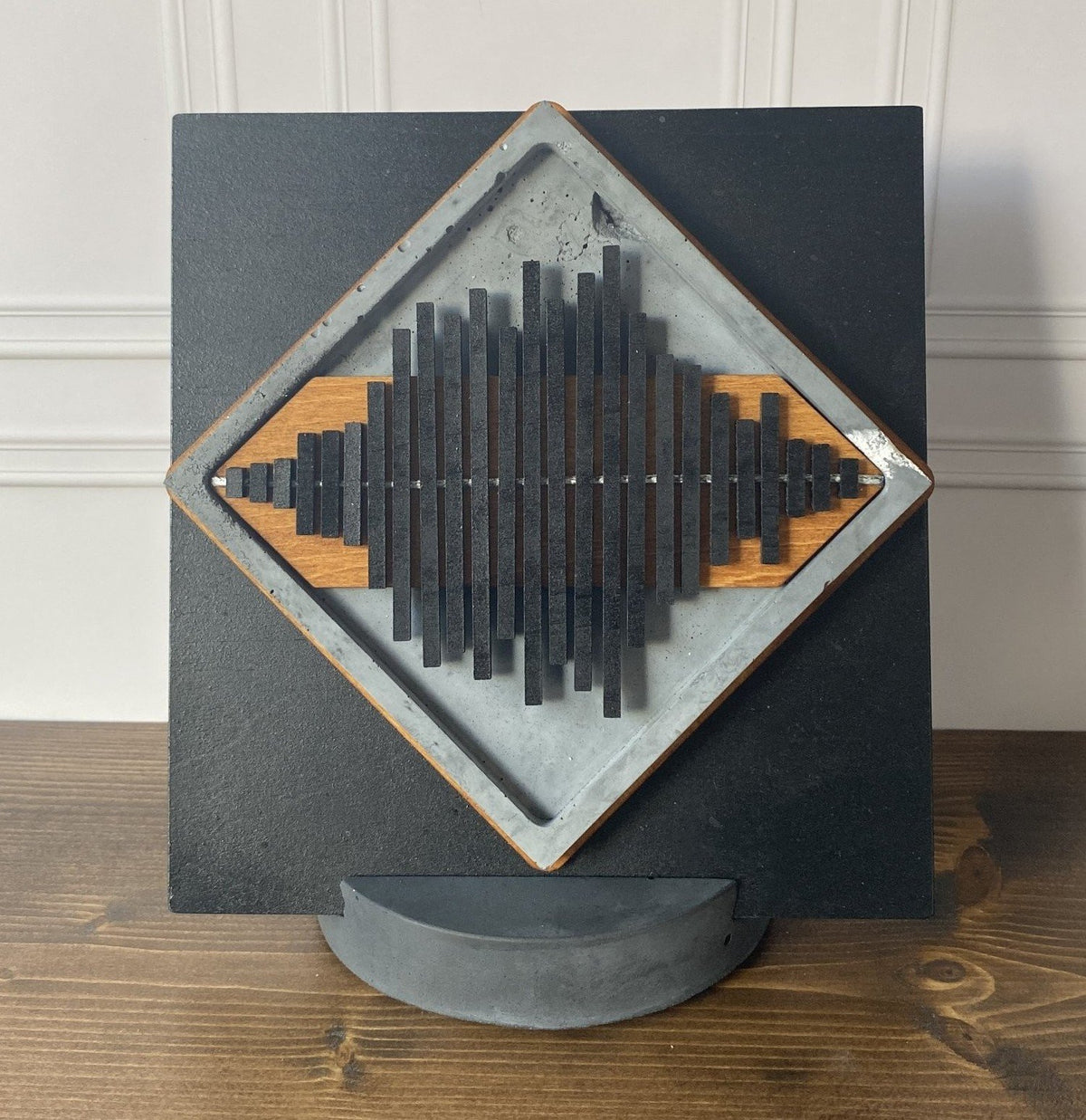 A square black wall-mounted art piece with a diamond-shaped inset featuring layered black and natural wood slats, displayed on a curved gray stand against a white paneled wall and wood tabletop.