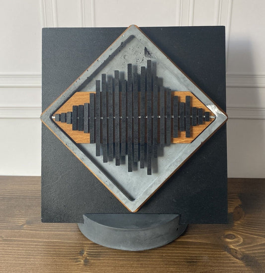 A square black wall-mounted art piece with a diamond-shaped inset featuring layered black and natural wood slats, displayed on a curved gray stand against a white paneled wall and wood tabletop.