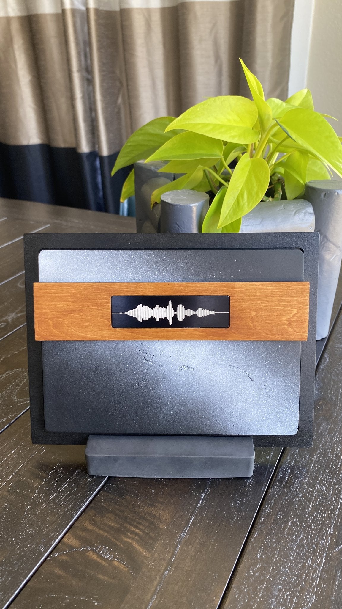 A modern soundwave-themed with a black frame, gray textured background, and a light wood accent band featuring a black soundwave emblem, displayed on a dark wooden table with a potted green plant in the background.