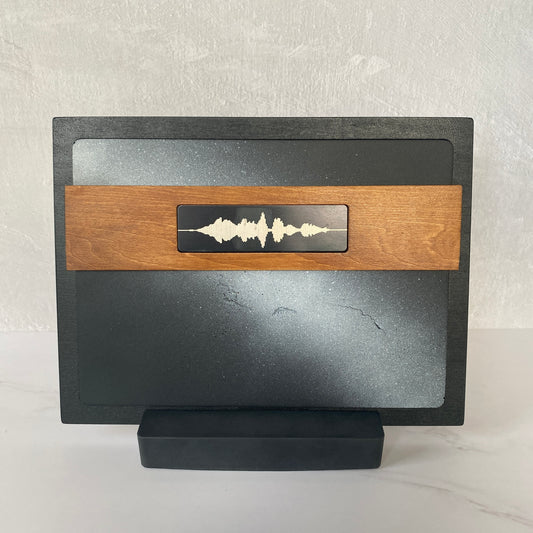 A modern soundwave-themed desk stand with a black frame, gray textured insert, and a light wood accent band featuring a black soundwave emblem, displayed on a dark wooden table.