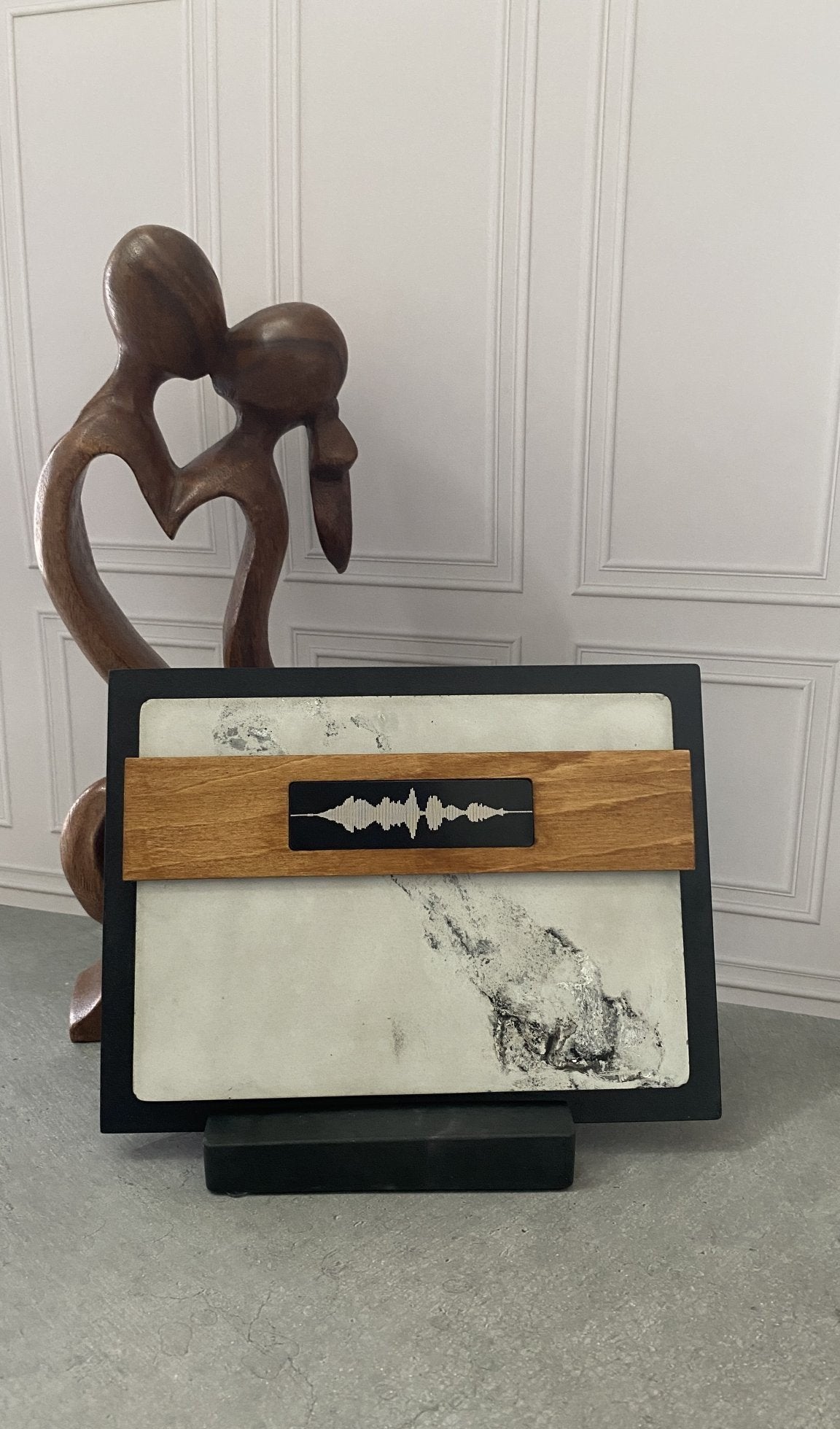 A rectangular black-framed wall art piece with a white marble lower section and a light wood horizontal band across the center, featuring a black inlay with a white soundwave design. The piece is displayed on a black stand in front of a white paneled wall, with a brown wooden abstract sculpture behind it.