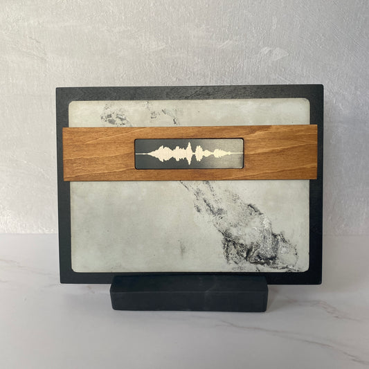 A rectangular decorative plaque or picture frame with a black border, light marble insert, and a horizontal wooden band across the center featuring a metallic soundwave design, displayed on a black stand against a light gray textured wall.
