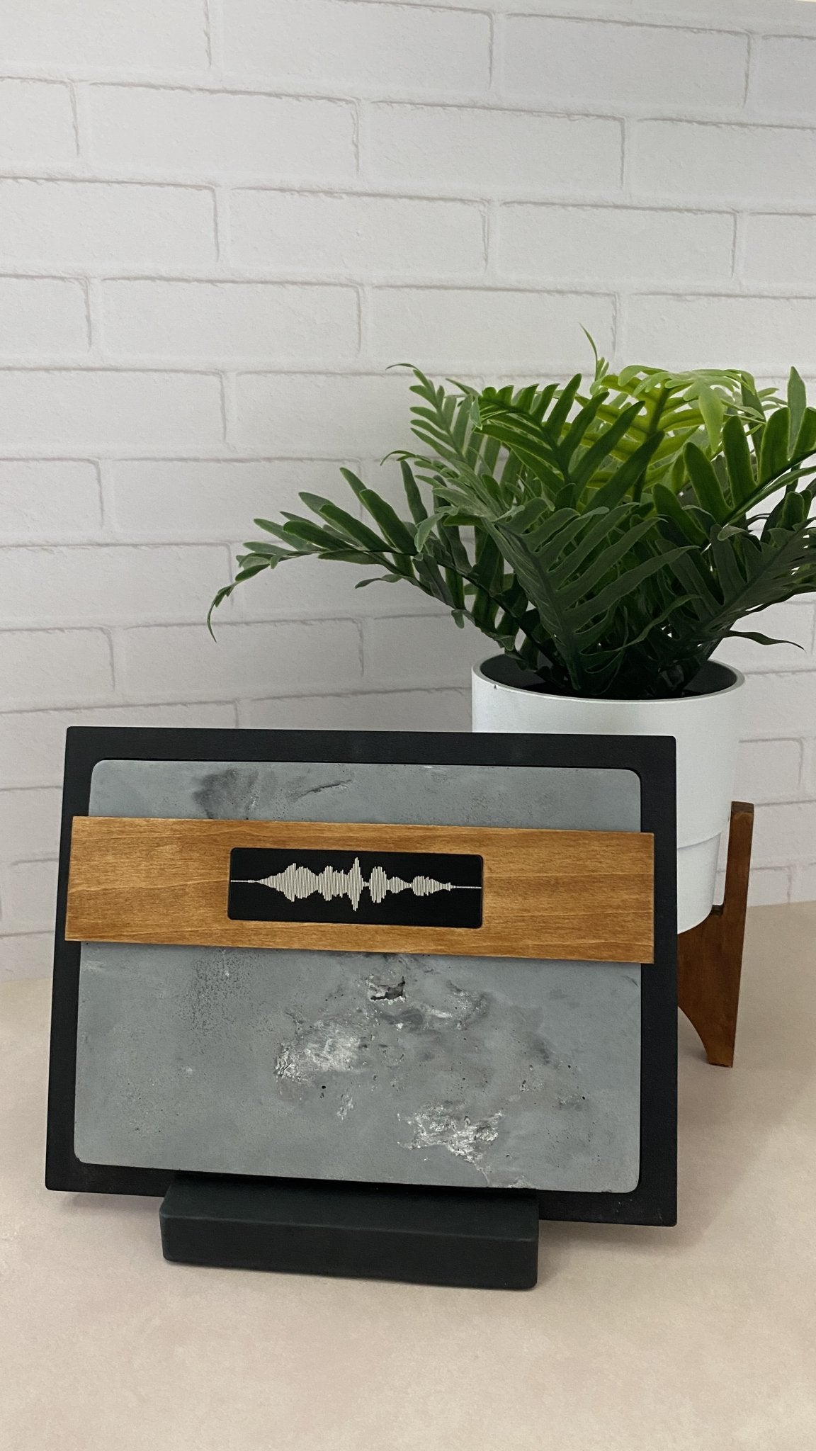 A square decorative board or tray with a gray stone-like center, black border, and a horizontal wooden band featuring a black rectangular inset with a white soundwave design. The board is displayed on a stand in front of a white brick wall, with a potted green plant behind it.