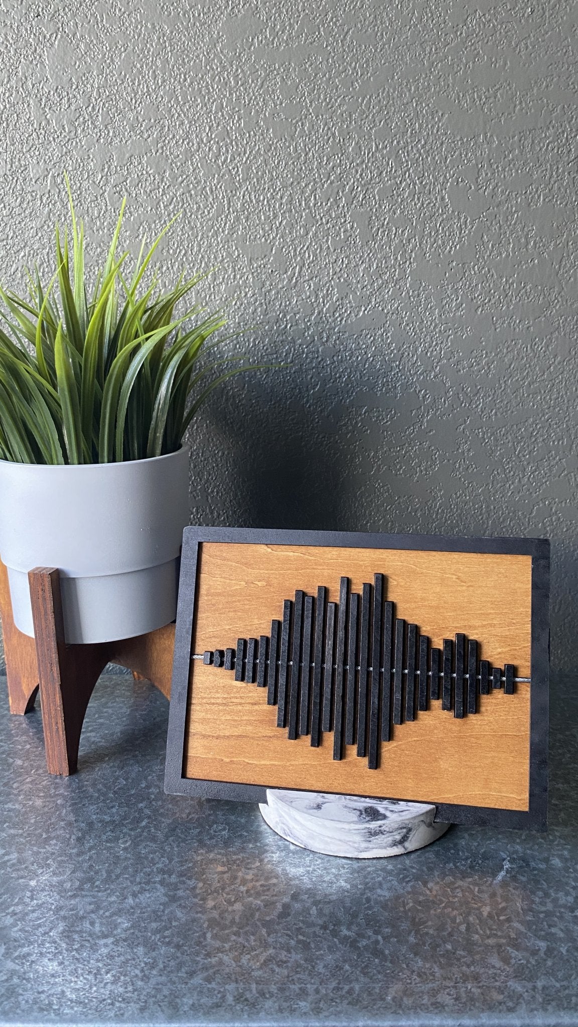 Stained wooden display piece with black sound wave and marbled stand with a green plant.