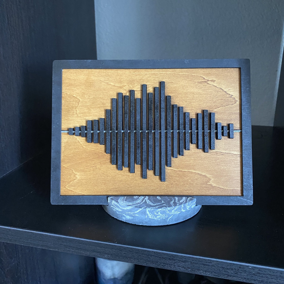 A rectangular wooden plaque with a dark soundwave design in the center, displayed on a round marble-like stand on a dark shelf.