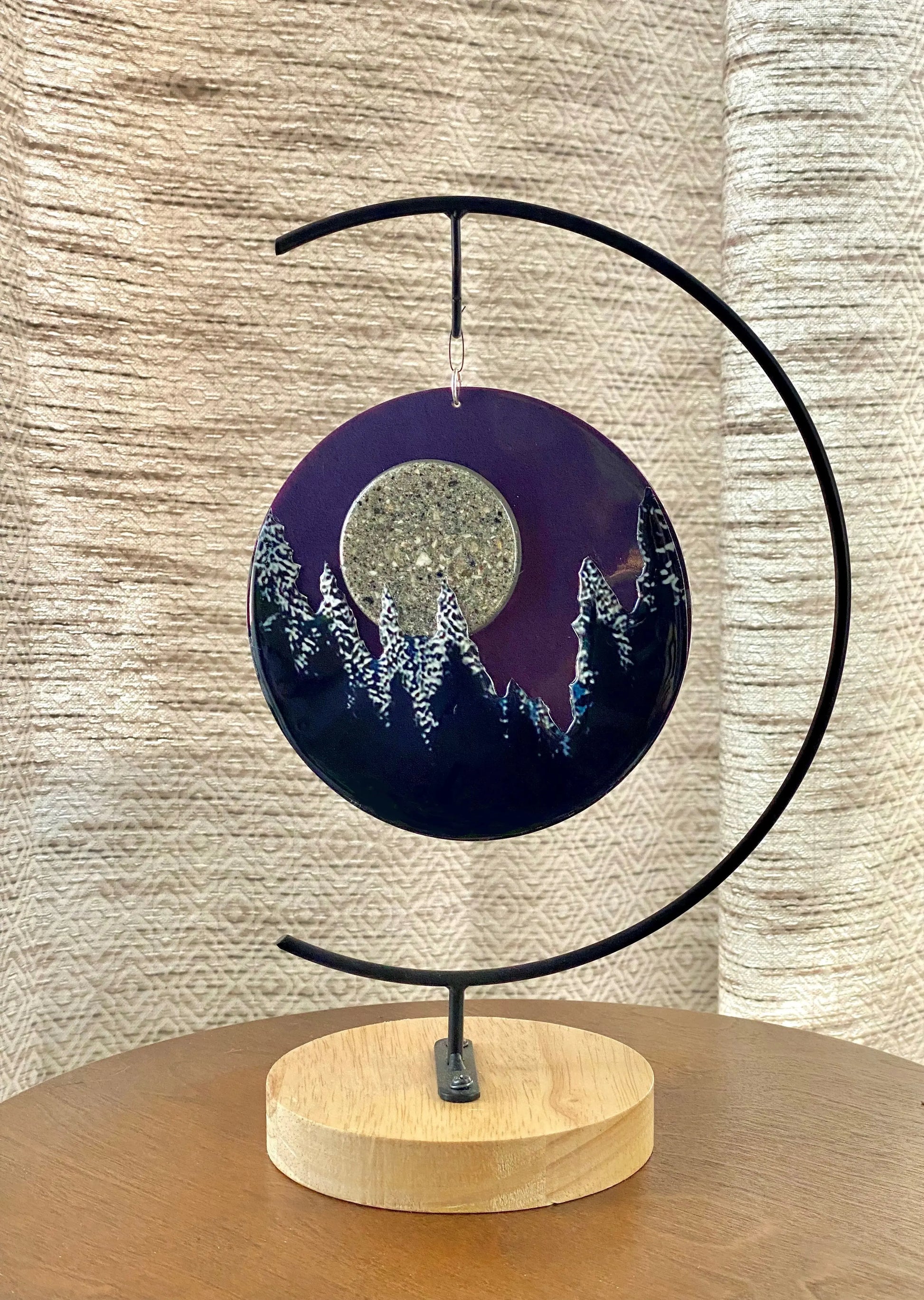 Decorative piece with a circular design featuring a moon and trees on a wooden base, against a textured beige wall.