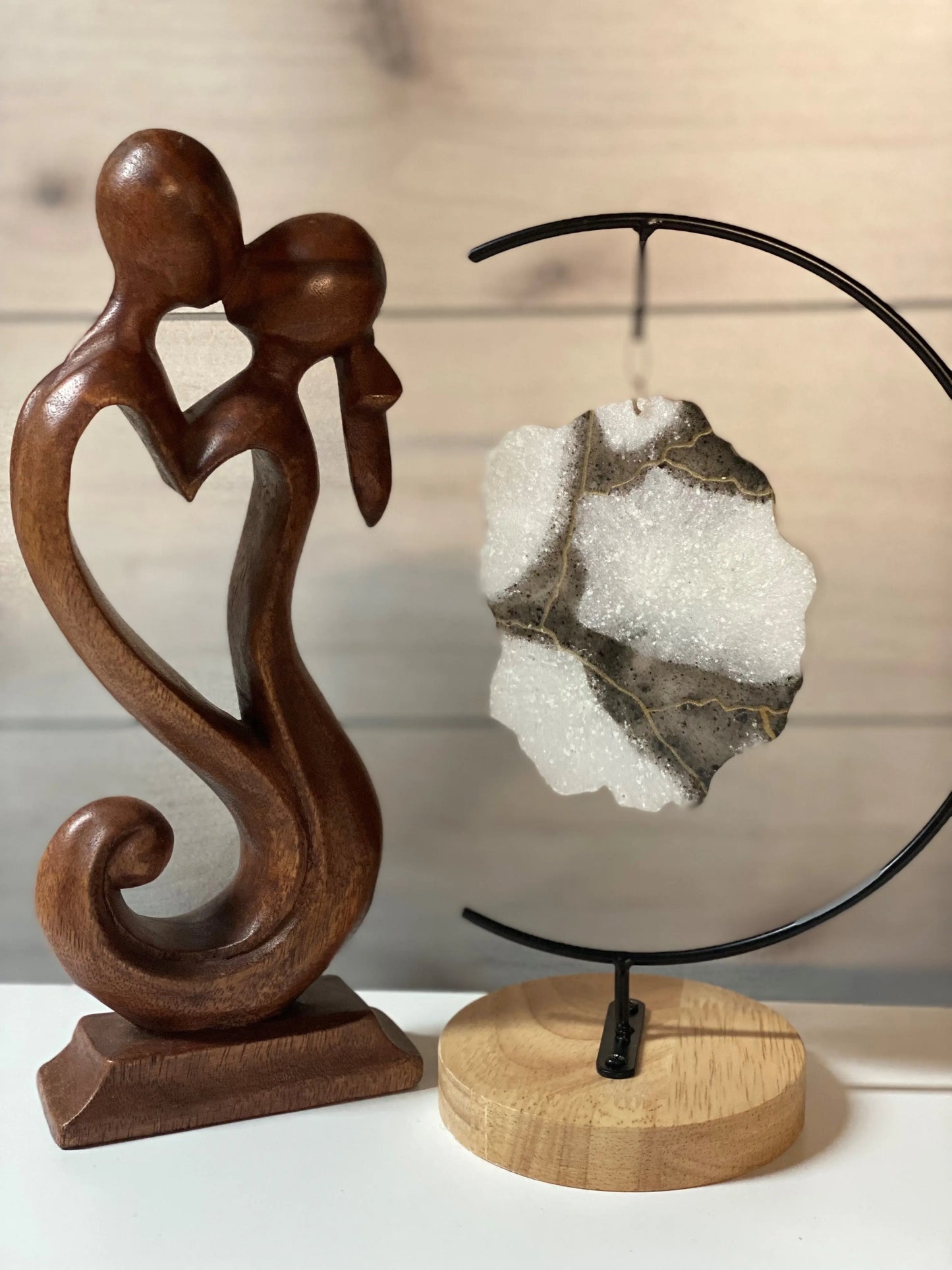 Wooden sculpture of two figures embracing on a white surface with a white-infused ash geode on a stand.