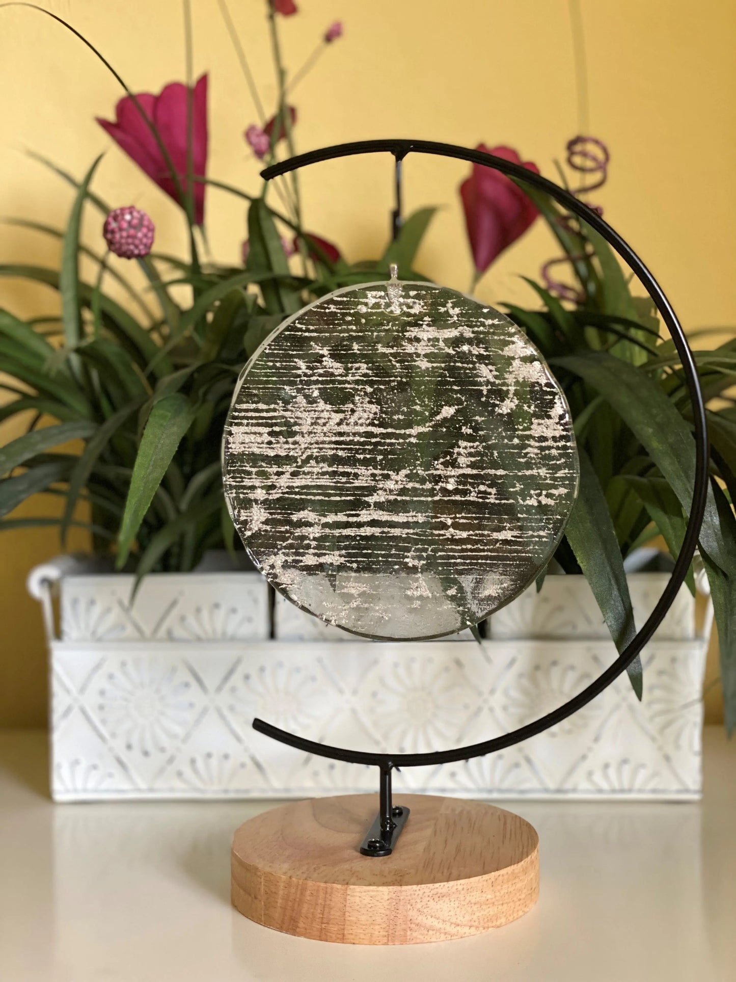 Decorative circular object infused with ash on a wooden stand with plants in the background