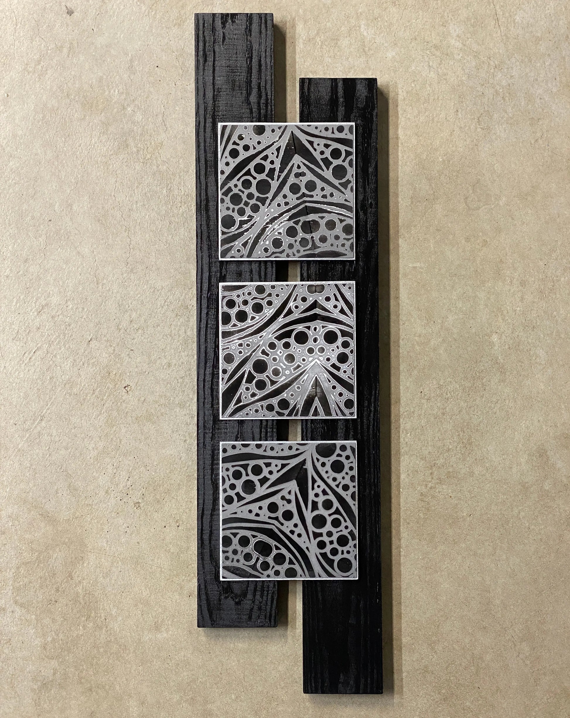 Vertical mixed‑media artwork featuring three etched metal tiles mounted on dark wooden planks, each with geometric patterns and circular cutouts.