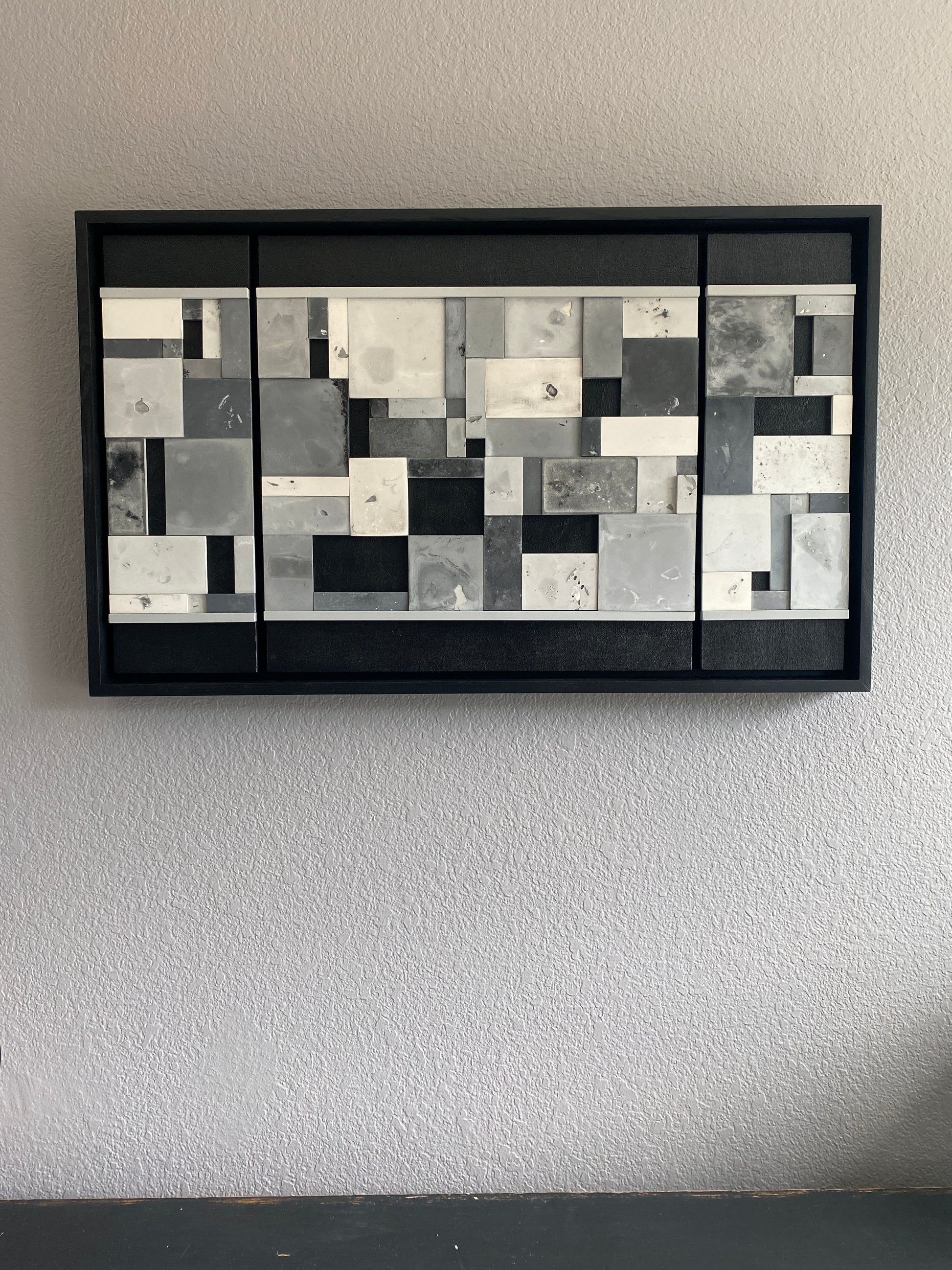 Abstract art piece with geometric shapes in black, white, and gray on a textured wall.