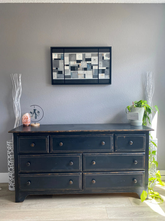 Black cabinet with multiple drawers against a gray wall, with decorative wall art in the center.