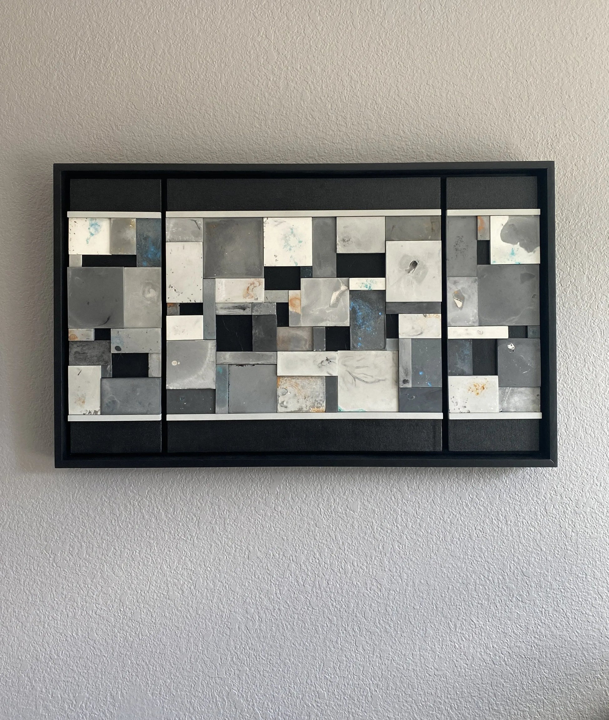 Abstract wall art with geometric shapes in a black frame on a plain wall.